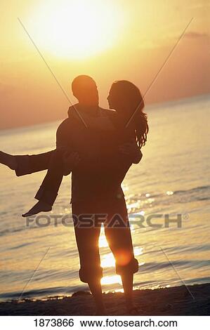 Stock Photograph - a man holding a woman on a beach in the sunset. Fotosearch