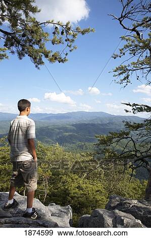 Stock Photo - a teenage boy stands on a rock cliff overlooking the mountains. Fotosearch