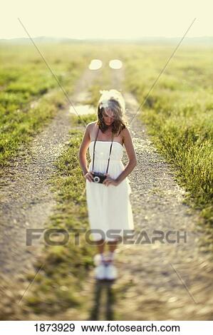 Stock Photo - a woman in a white dress standing on a dirt road with a camera around her neck. Fotosearch