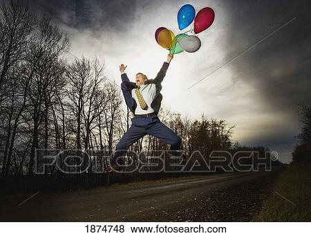 Stock Photo - alberta, canada; a businessman jumping in the air on the side of the road holding balloons. Fotosearch