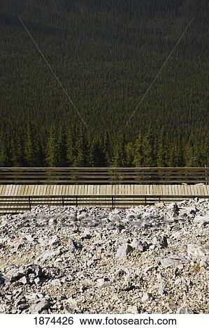 Stock Photograph - banff, alberta, canada; a wooden boardwalk with railings on a mountain with rock on one side and forest on the other. Fotosearch