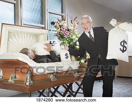 Stock Image - edmonton, alberta, canada; a happy man holds bags of money beside a deceased man in a coffin. Fotosearch