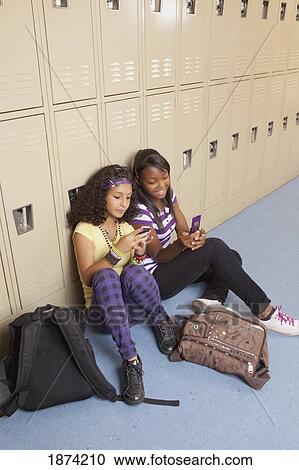 Stock Image - fort lauderdale, florida, united states of america; two teenage girls using their cell phones while sitting in a school hallway. Fotosearch