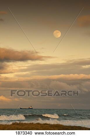 Stock Image - saltburn, teesside, england; a cargo ship traveling on the ocean with the moon in the sky. Fotosearch