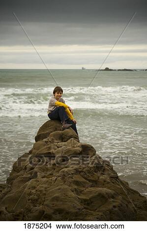 Picture - a boy sits on a rock at the water's edge. Fotosearch
