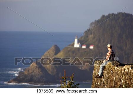 Stock Photo - oregon, united states of america; a man sitting on a cliff looking out over the pacific ocean with heceta head lighthouse in the background. Fotosearch