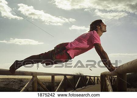 Stock Image - tarifa, costa de la luz, cadiz, andalusia, spain; a man stretching on the wooden railing of a boardwalk at bunker beach. Fotosearch