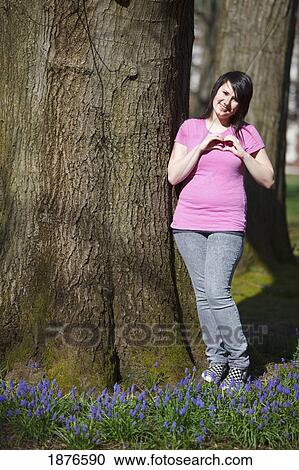 Stock Image - Portland, Oregon, United States Of America; A Teenage Girl Standing By A Tree And Making A Heart Shape With Her Hands Against Her Chest. Fotosearch