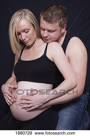 Stock Photo - a man embracing a pregnant woman as they touch her bare belly. Fotosearch