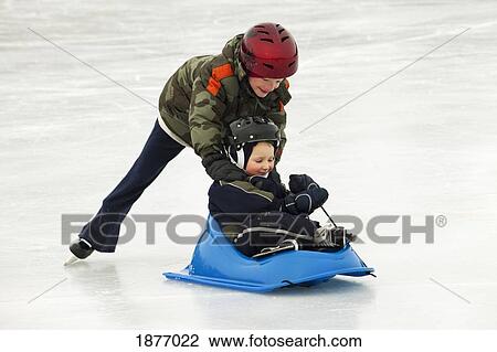 Stock Image - edmonton, alberta, canada; a boy pushing a younger boy in a sled on an outdoor ice rink. Fotosearch