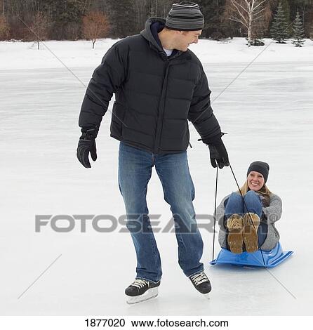 Stock Image - edmonton, alberta, canada; a man pulling a woman on a small sled across the ice on an outdoor rink. Fotosearch