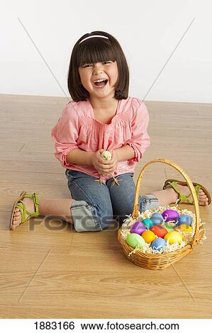 Stock Photograph - A Girl Holding A Chick And Sitting With A Basket Full Of Colorful Easter Eggs. Fotosearch
