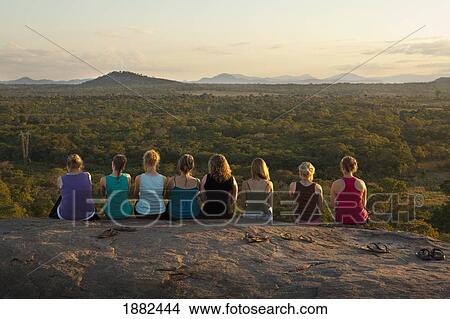 Picture - A Row Of Young Women Sitting On A Rock Ledge Viewing The African Landscape; Manica, Mozambique, Africa. Fotosearch