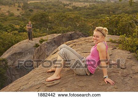 Stock Image - A Young Woman Sits On A Rock With The African Landscape In The Background; Manica, Mozambique, Africa. Fotosearch