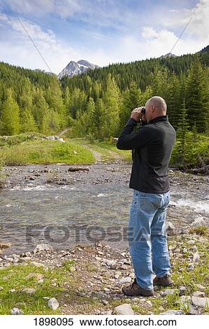Stock Photography - a man looking through binoculars as he hikes through the mountains; fernie, british columbia, canada. Fotosearch