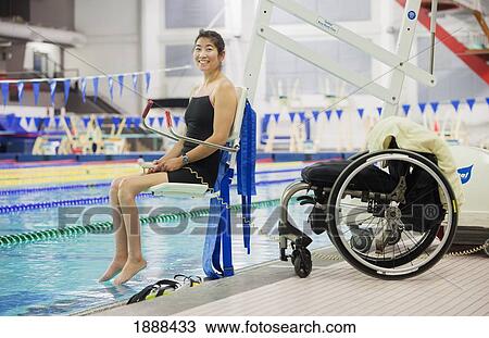 Stock Image - a paraplegic woman enters a swimming pool on a lift with her wheelchair at the edge of the pool; edmonton, alberta, canada. Fotosearch