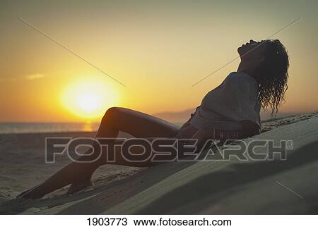 Stock Image - a woman wearing a bathing suit lies on the sand on los lances beach at sunset; tarifa, cadiz, andalusia, spain. Fotosearch