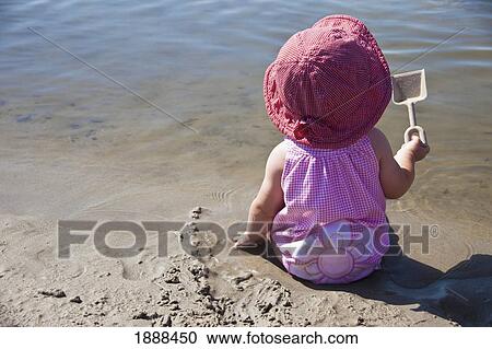 Stock Image - a young girl sits in the wet sand at the water's edge holding a shovel; county of st. paul, alberta, canada. Fotosearch