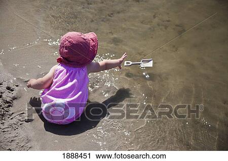 Stock Image - a young girl sits in the wet sand at the water's edge reaching for her floating shovel; county of st. paul, alberta, canada. Fotosearch