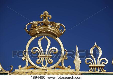 Stock Image - gold crown and fleur-de-lis on the grand trianon gates against a blue sky in the gardens of the palace of versailles; paris, france. Fotosearch