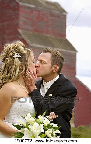 Stock Image - groom embracing his bride as he kisses her in front of an old country grain elevator; st. albert, alberta, canada. Fotosearch