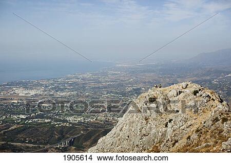 Picture - two mountain climbers sitting on a peak overlooking the city; marbella, malaga, andalusia, spain. Fotosearch
