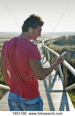 Stock Photography - a man listening to music with earbuds as he walks down a wooden promenade towards dos mares beach; tarifa, cadiz, andalusia, spain. Fotosearch
