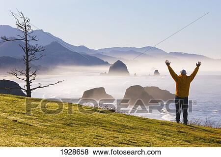 person with raised arms on hillside at sunrise and morning fog add beauty to cannon beach and haystack rock from ecola state park; oregon, united states of america View Large Photo Image Stock Photo - person with raised arms on hillside at sunrise and morning fog add beauty to cannon beach and haystack rock from ecola state park; oregon, united states of america. Fotosearch