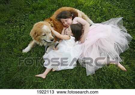 Stock Image - two sisters dressed in princess dresses hug their rescue dog, bo, while laying on the lawn; duncan, british columbia, canada. Fotosearch