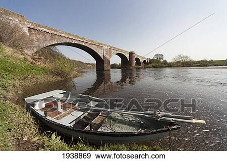 Stock Photo - A Boat Along The Shoreline Beside A Bridge In River Tweed; Scottish Borders Scotland. Fotosearch