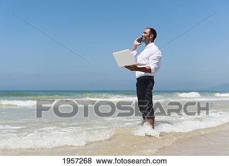 a businessman holds his laptop computer and talks on the cell phone while standing in the surf on the beach; tarifa cadiz andalusia spain View Large Photo Image Stock Photo - a businessman holds his laptop computer and talks on the cell phone while standing in the surf on the beach; tarifa cadiz andalusia spain. Fotosearch