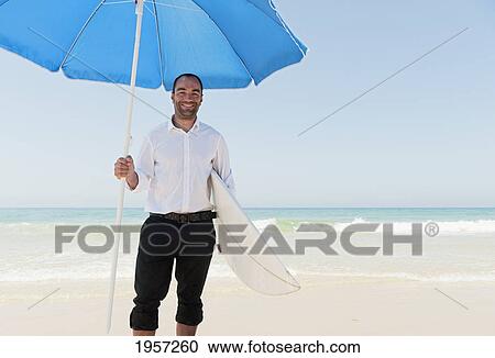 Stock Image - a businessman on the beach holding a beach umbrella and surfboard; tarifa cadiz andalusia spain. Fotosearch