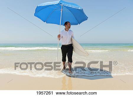 Stock Image - a businessman on the beach holding a beach umbrella and surfboard; tarifa cadiz andalusia spain. Fotosearch