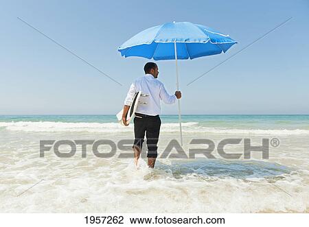 Stock Image - a businessman on the beach holding a beach umbrella and surfboard; tarifa cadiz andalusia spain. Fotosearch