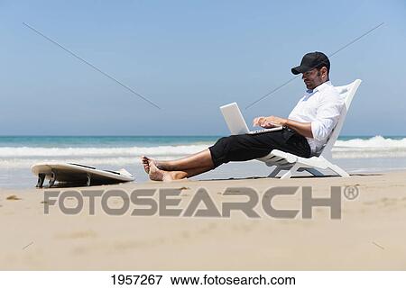 Stock Photo - a businessman sits on a beach chair on the beach working on a laptop computer with his surfboard sitting at his feet; tarifa cadiz andalusia spain. Fotosearch