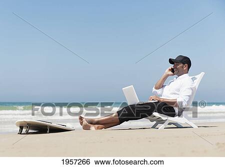 a businessman sits on a beach chair on the beach working on a laptop computer and talking on the phone with his surfboard sitting at his feet; tarifa cadiz andalusia spain View Large Photo Image Stock Photograph - a businessman sits on a beach chair on the beach working on a laptop computer and talking on the phone with his surfboard sitting at his feet; tarifa cadiz andalusia spain. Fotosearch