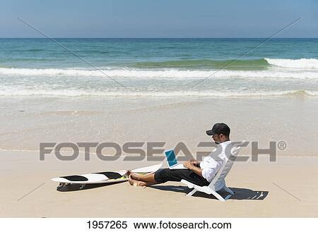 Stock Photography - a businessman sits on a beach chair on the beach working on a laptop computer with his surfboard sitting at his feet; tarifa cadiz andalusia spain. Fotosearch