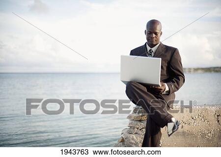 Stock Image - A Businessman Using A Laptop Computer On The Water's Edge; South Caicos Turks And Caicos Islands. Fotosearch
