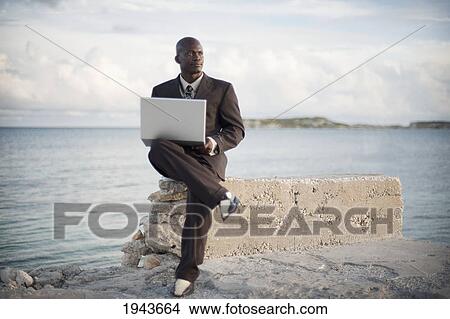 Picture - A Businessman With A Laptop Computer Sitting On A Rock Ledge At The Edge Of The Water; South Caicos Turks And Caicos Islands. Fotosearch