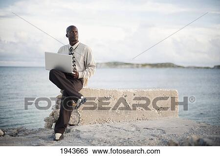 Stock Photography - A Businessman With A Laptop Computer Sitting On A Rock Ledge At The Edge Of The Water; South Caicos Turks And Caicos Islands. Fotosearch
