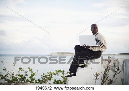 Stock Image - A Businessman Working On A Laptop Computer By The Water's Edge; South Caicos Turks And Caicos Islands. Fotosearch