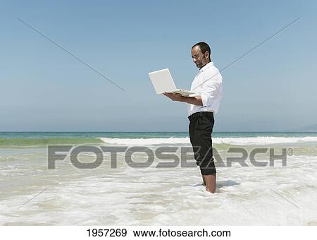 Stock Photo - a businessman works on his laptop computer while standing in the surf on the beach; tarifa cadiz andalusia spain. Fotosearch
