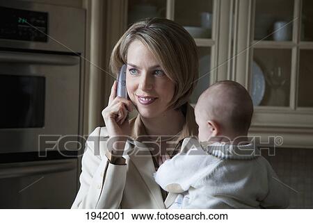 Stock Image - A Businesswoman Holding Her Baby And Talking On The Phone In The Kitchen At Home; Jordan Ontario Canada. Fotosearch