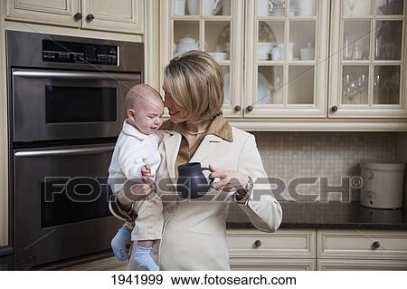 A Businesswoman Holding Her Baby In The Kitchen At Home; Jordan Ontario Canada View Large Photo Image Stock Photo - A Businesswoman Holding Her Baby In The Kitchen At Home; Jordan Ontario Canada. Fotosearch