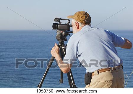 Stock Image - a man operating a video recorder mounted on a tripod looking out over the water; malaga province spain. Fotosearch