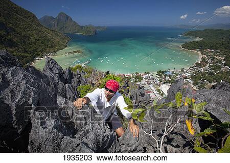 Stock Image - A Man Rock Climbs On Top Of Sharp Limestone Spires Overlooking The Village Of El Nido; El Nido, Bacuit Archipelago, Palawan, Philippines. Fotosearch