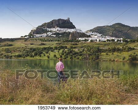 Stock Photo - a man standing at water's edge looking at zahara de la sierra as seen over the zahara dam; zahara de la sierra cadiz andalusia spain. Fotosearch