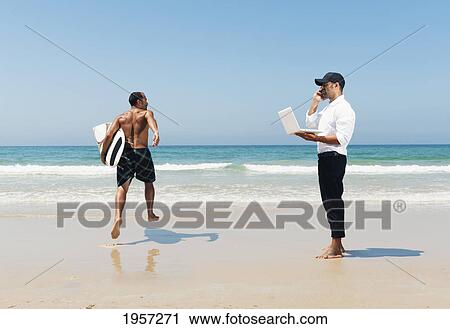 Stock Image - a man standing on a beach representing himself with a surfboard on vacation and working as a businessman; tarifa cadiz andalusia spain. Fotosearch