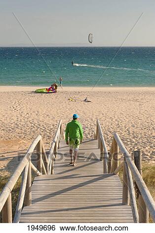 Stock Photograph - a man walks down a wooden boardwalk to valdevaqueros beach; tarifa cadiz andalusia spain. Fotosearch
