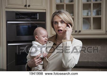 Stock Photo - A Mother Holding Her Baby In The Kitchen With A Worried Look While Talking On The Phone; Jordan Ontario Canada. Fotosearch